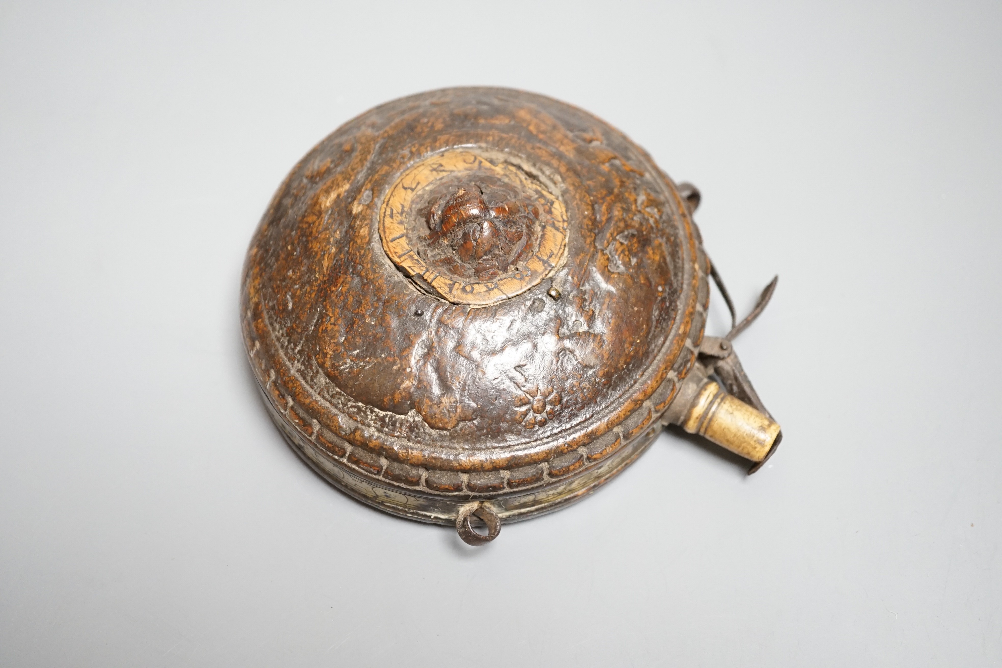 A circular wooden powder flask 17th century, carved with bear, hound and fox, and lion’s mask in low relief surrounded by game counter, engraved brass border, iron lever charger, turned bone nozzle. Diameter 11cms